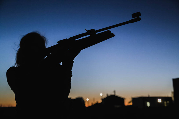 silhouette of a girl who at night on the background of houses aim with a rifle - Photo, Image