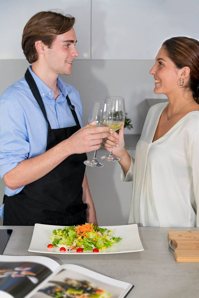 Jeune couple souriant portant un toast avec des verres de vin blanc à côté d'un comptoir de cuisine avec une salade et un livre de recettes flou. Cuisine à la maison et concept amusant
. - Photo, image