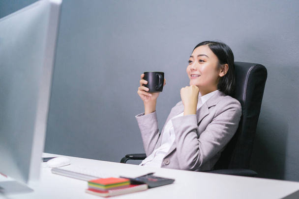 Ευτυχισμένη Ασιάτισσα επιχειρηματίας που πίνει καφέ στο γραφείο της και χαμογελάει.. - Φωτογραφία, εικόνα