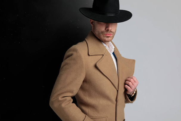 πλαϊνή άποψη του μυστηριώδους νεαρού άνδρα με μακρύ παλτό φορώντας καπέλο και ποζάροντας με τρόπο μόδας, στέκεται σε μαύρο γκρι φόντο - Φωτογραφία, εικόνα
