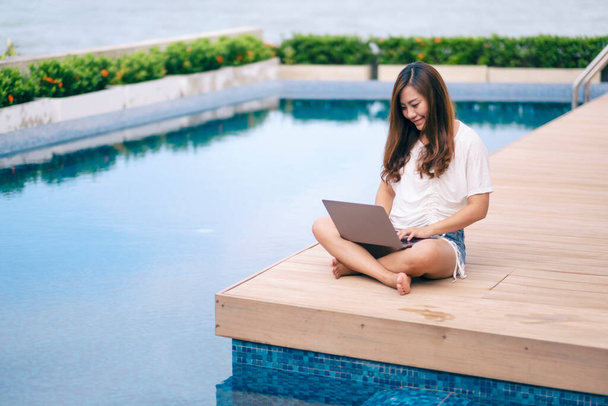 Een mooie aziatische vrouw met behulp van en typen op laptop computer tijdens het zitten bij het zwembad - Foto, afbeelding