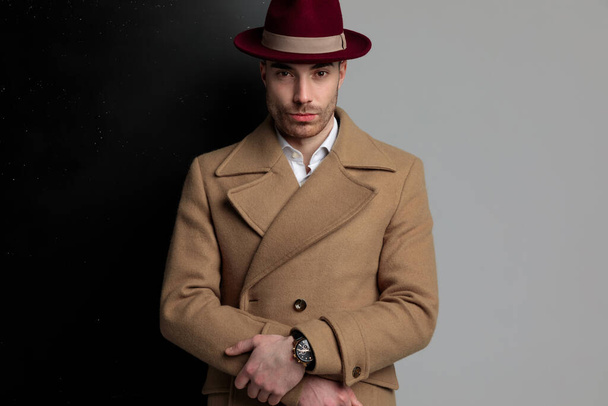 young fashion guy wearing long coat and hat, smiling and holding hands in a fashion pose, standing on black grey background - Fotografie, Obrázek