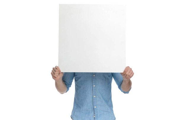 young casual guy covering face with empty board and standing isolated on white background, portrait - 写真・画像
