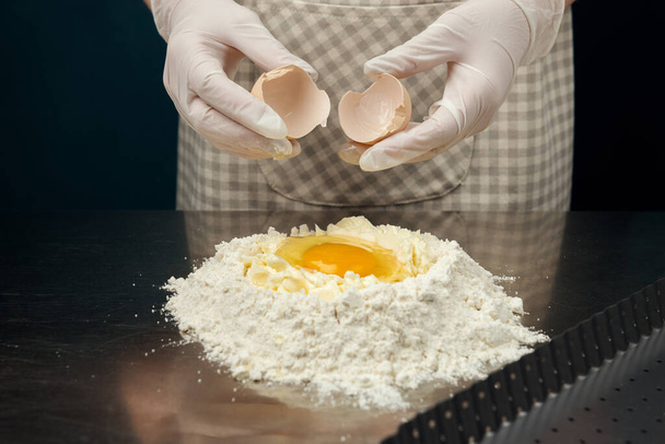 Kucharz rozkłada jajko na mieszankę mąki i masła na stole kuchennym ze stali nierdzewnej. Bezpieczny proces gotowania tarty, ciasteczek lub piekarni. Ręce rękawiczki. - Zdjęcie, obraz
