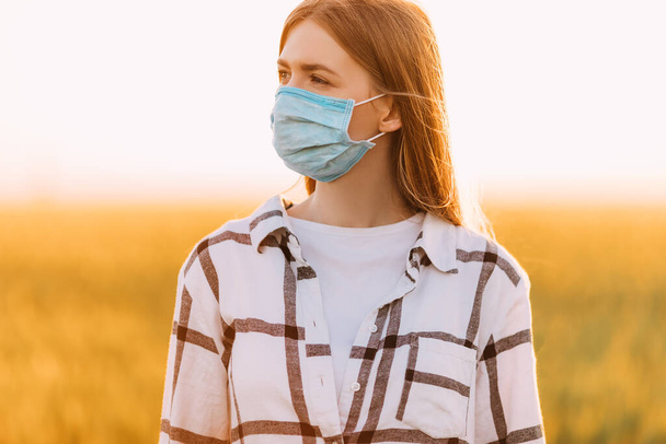beautiful woman in a checked shirt and a protective medical mask on her face stands in a wheat field at sunset. Quarantine, coronavirus, crop - Fotó, kép
