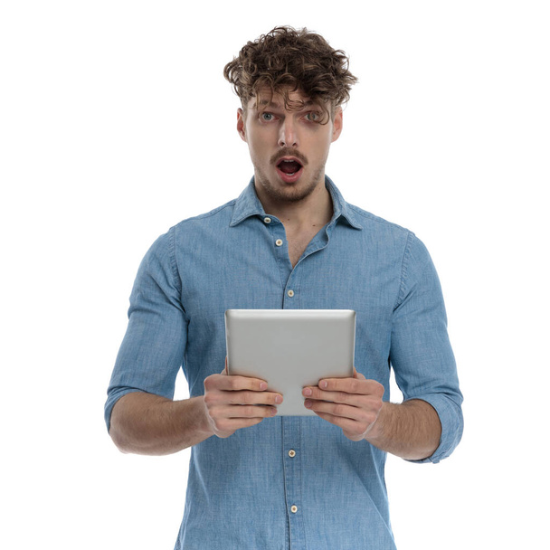 šokovaný mladý ležérní model v džínové košili drží tablet a stojí izolované na bílém pozadí, portrét - Fotografie, Obrázek