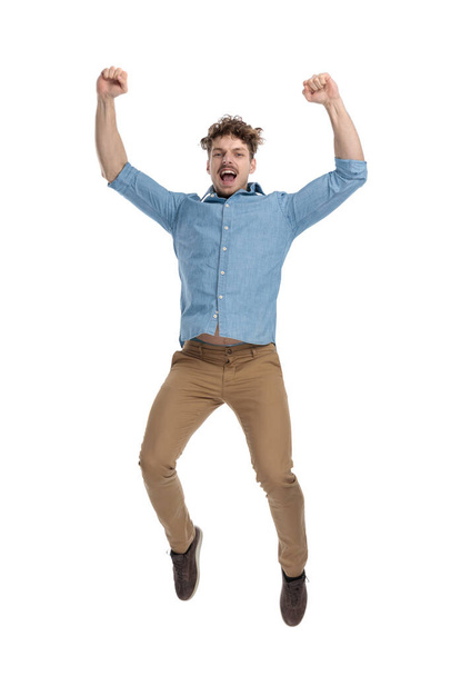excited young casual guy in denim shirt holding arms in the air, jumping and celebrating victory isolated on white background, full body - Photo, Image