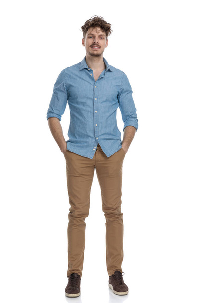 cara casual atraente na camisa jeans de mãos dadas em bolsos e sorrindo isolado no fundo branco, corpo inteiro
 - Foto, Imagem