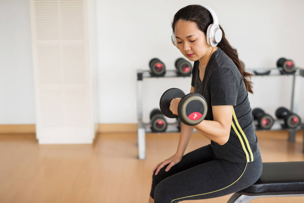 Fitness Mujer asiática sentarse en la silla para preformar el ejercicio de la mancuerna bíceps en el gimnasio. Culturismo y deporte concepto de estilo de vida saludable. Chica china entrenando sola. Distanciamiento social, nueva normalidad
. - Foto, Imagen