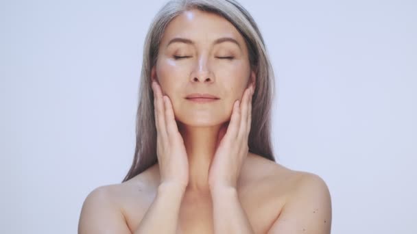 A happy pleased old mature half-naked woman with long gray hair is touching her ideal skin isolated over white background in studio - Záběry, video