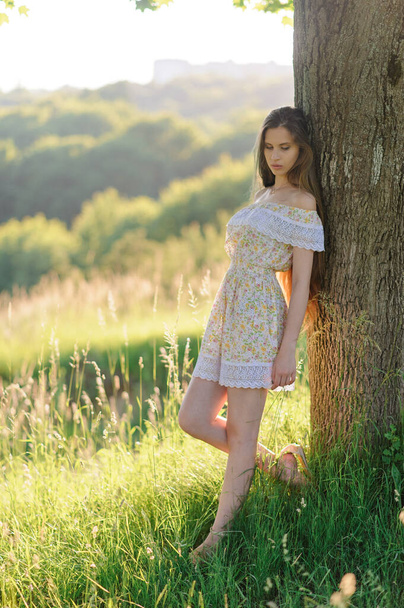 Retrato de una joven hermosa en un vestido de sol. Sesión de fotos de verano en el parque al atardecer. Chica se inclina contra un árbol
. - Foto, imagen