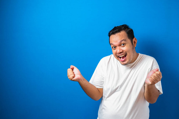 Portré egy vicces kövér ázsiai férfi fehér pólóban mosolyog és táncol boldogan, örömteli kifejező ünneplő jó hír győzelem győztes siker gesztus ellen kék háttér - Fotó, kép