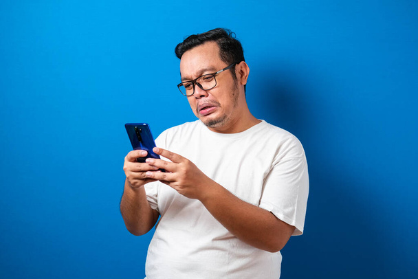 Толстый азиат в белой футболке выглядит грустным, читая онлайн новости со своего сотового. мужчина показывает разочарованный жест, вытирая слезы с лица
 - Фото, изображение