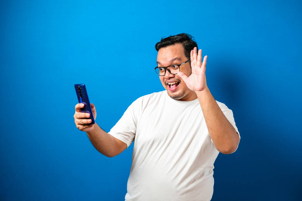 Portrait studio d'un jeune homme d'Asie de l'Est en t-shirt blanc faisant un appel vidéo et prendre selfie, sur fond bleu
 - Photo, image