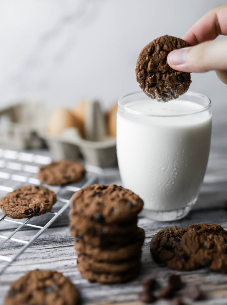 a hand dipping cookie in a tall glass of milk with a stack of homemade chocolate chip cookies - Foto, Imagem