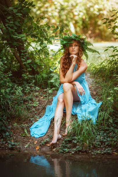 egy gyönyörű fiatal nő vörös hajjal, koszorúval, virágokkal és páfránnyal az erdőben ünnepli a napforduló napját, nyár közepén.. - Fotó, kép