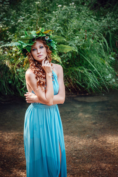 eine schöne junge Frau mit roten Haaren und einem Kranz aus Blumen und Farn im Wald feiert den Sonnwendtag, mitten im Sommer. - Foto, Bild