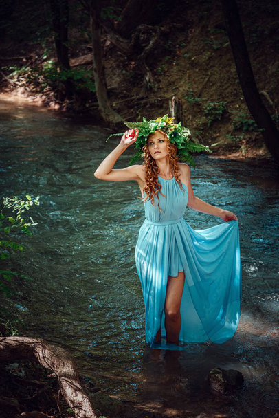Μια όμορφη νεαρή γυναίκα με κόκκινα μαλλιά και στεφάνι από λουλούδια και φτέρη στο δάσος γιορτάζει την Ημέρα του Ηλιοστασίου, στα μέσα του καλοκαιριού.. - Φωτογραφία, εικόνα