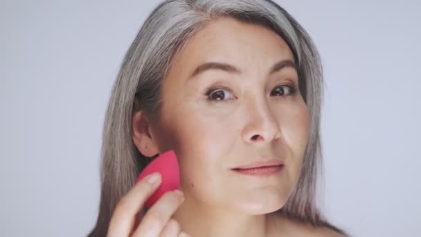 A beautiful old mature half-naked woman with long gray hair is doing a make-up using a pink sponge isolated over white background in studio - Imágenes, Vídeo