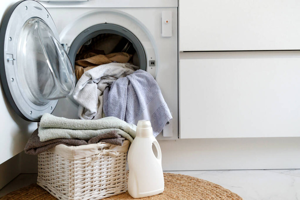 Carregar uma máquina de lavar e lavar roupa. Uma cesta de roupa e gel de lavanderia na frente de uma máquina de lavar roupa doméstica embutida na cozinha
. - Foto, Imagem