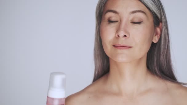 A smiling old mature half-naked woman with long gray hair is showing a care-cosmetics isolated over white background in studio - Filmati, video