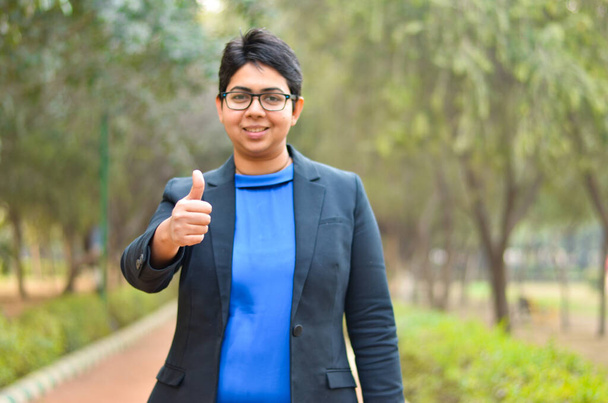 Портрет упевненої молодої індійської корпоративної професійної жінки з коротким волоссям з пальцями вгору знак на відкритому повітрі, одягнений в чорний бізнес / формальний костюм - Фото, зображення