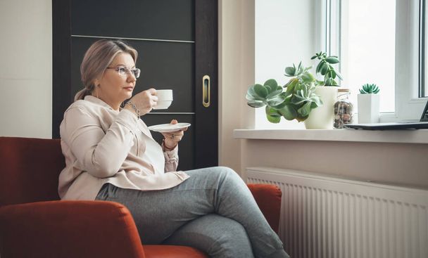 Mulher caucasiana sênior com óculos sentados em poltrona e bebendo um café enquanto usava óculos e assistindo a uma lição no laptop
 - Foto, Imagem