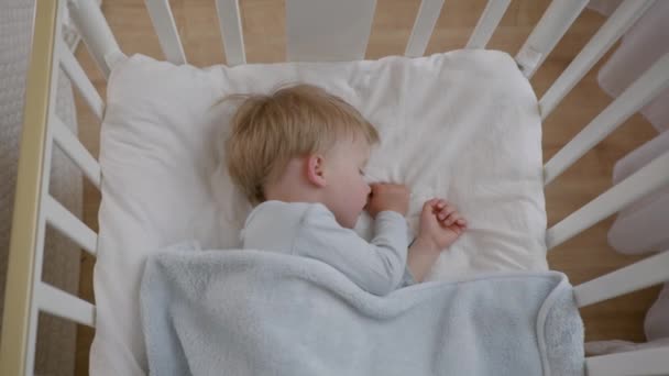 ベビーベッドに毛布で覆われた柔らかい快適な枕の上で、ハンサムな子供は昼間に健全に眠ります - 映像、動画