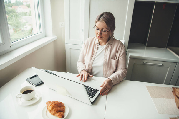 Donna caucasica anziana che lavora a distanza al computer portatile da casa mentre beve un caffè con croissant
 - Foto, immagini