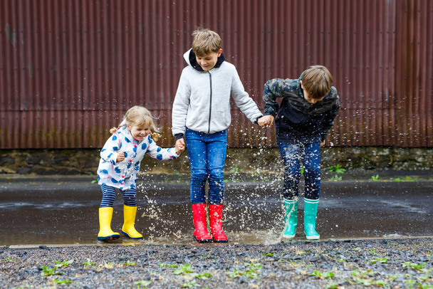 Tres niños, una niña pequeña y dos niños con botas de lluvia rojas, amarillas y verdes y caminando durante el aguanieve. Felices hermanos saltando al charco. Divertirse al aire libre, familia activa
 - Foto, imagen
