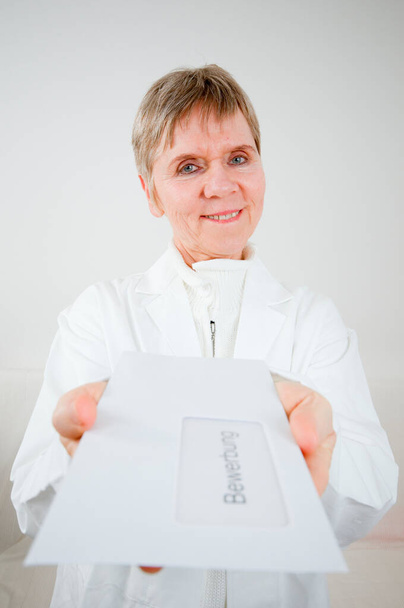 Frontal torso portrait of an older woman in a white coat in front of an envelope with the words "Application" towards the camera, holding a white background. - Photo, Image