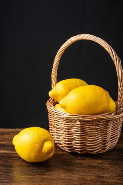 Nahaufnahme von Zitronen im Korb mit Griff und ganzer Zitrone auf Holztisch und schwarzem Hintergrund, vertikal mit Kopierraum - Foto, Bild