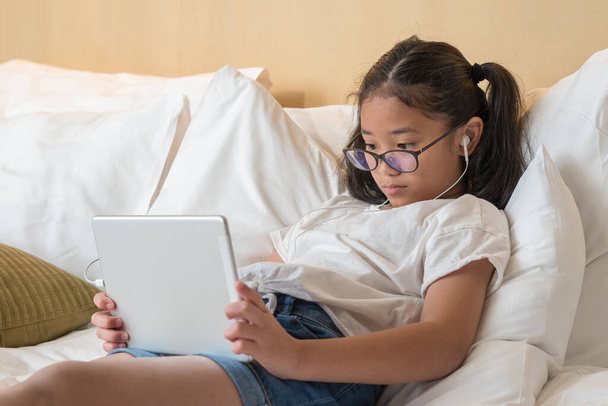Escuela chica asiática niño usando ipad tableta de computadora digital en la cama en casa para la educación e-learning lectura de libros electrónicos y jugar juego para la diversión y la relajación a través de Internet
 - Foto, Imagen