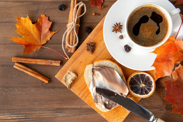 Cup of black coffee with a saucer, orange autumn maple leaves, cinnamon sticks, anise stars ,  bread with creamy chocolate paste on a brown wooden background top view - Fotografie, Obrázek