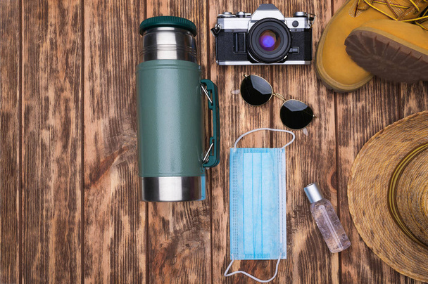 Travel accessories set on wooden background: face mask, antibacterial gel, hiking leather boots, straw hat, stainless steel thermos, vintage camera and sunglasses. Top view point. - Photo, Image