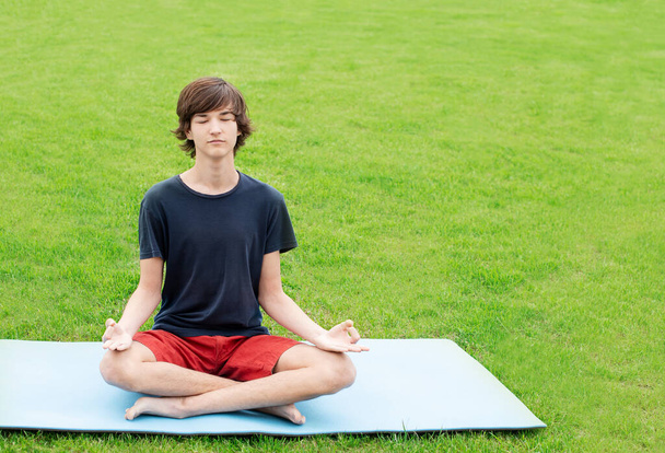 Un joven con el pelo largo hace yoga al aire libre. Un adolescente se sienta en una posición de loto en la hierba verde. Copiar espacio. Lugar para el texto
. - Foto, imagen
