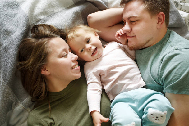 Top view of happy family in bed in the morning in casual clothes, young 30s parents, mother father and baby child lying and smiling - Valokuva, kuva
