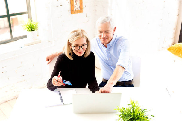 High angle shot of happy senior couple doing home finances. Attractive elderly woman holding bank card in her hand while senior man sitting behind their laptop and showing something.  - Foto, Bild