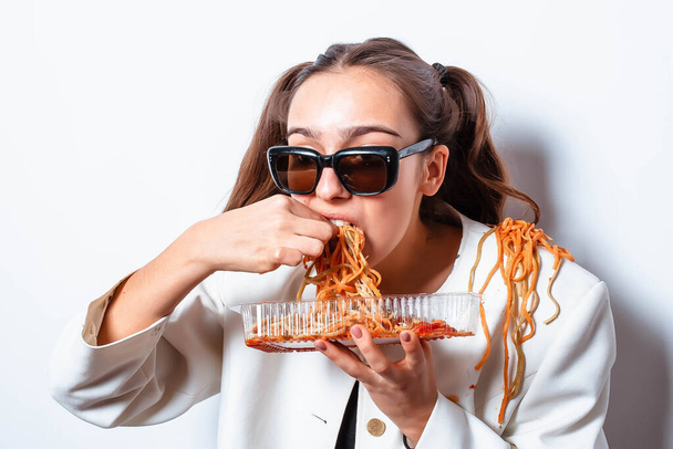 Krullend meisje in kleding en een bord met pasta en een vork eet wild pasta met ketchup, staande in glazen op een witte achtergrond. Meisje dat spaghetti eet. Meisje met een bord pasta. - Foto, afbeelding