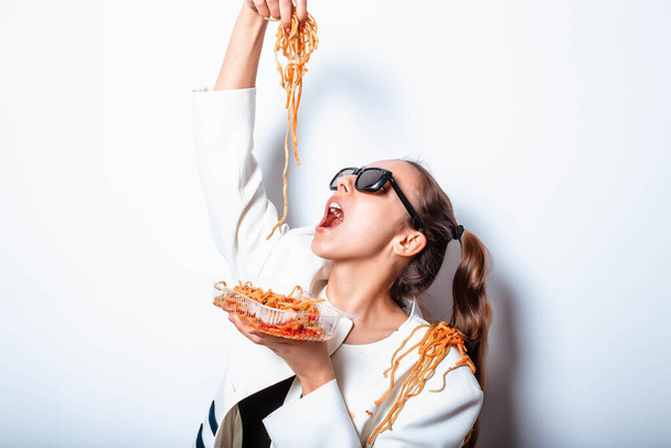 Szexi csábító provokatív lány ruhákban és egy tányér tésztával és egy villával vadul eszik tésztát ketchuppal, szemüvegben áll fehér háttérrel. Spagettit evő lány. - Fotó, kép