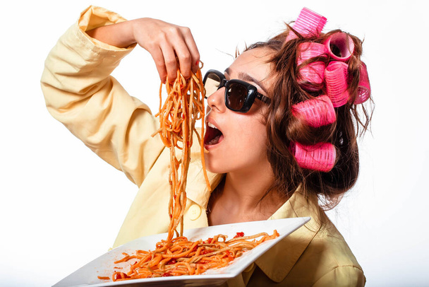 Curly girl in clothes and a plate with pasta and a fork wildly eats pasta with ketchup, standing in glasses on a white background. Girl eating spaghetti. Girl holding a plate of pasta. - Foto, immagini