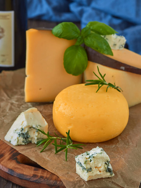 Various kinds of cheese and herbs on a rustic wooden table, closeup, wine in the background. Homemade cheeses on paper lining. Traditional slices of French and Italian cheese handmade - Photo, Image