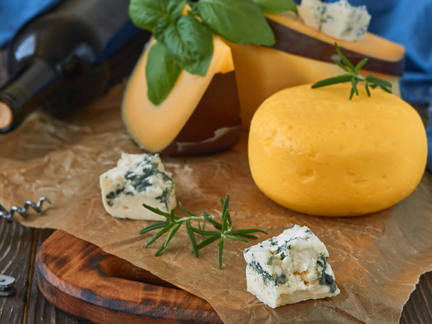 Fresh cheeses and herbs on a rustic wooden table, closeup, wine in the background. Homemade cheeses on paper lining. - Photo, Image