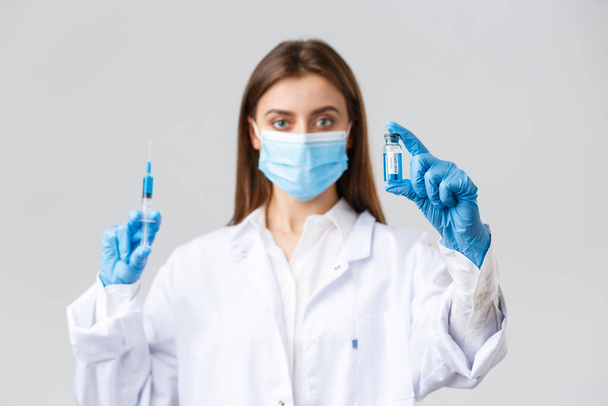 Covid-19, preventing virus, healthcare workers and quarantine concept. Doctor in medical mask, rubber gloves and scrubs showing ampoule with coronavirus vaccine, hold syringe, making patient a shot - Foto, imagen