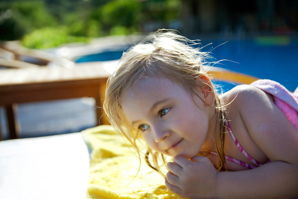 Little blond girl resting near the swimming pool. Outdoor and sunny holiday mood, Little cute blonde girl sunbathing in a lounge chair near the pool. - Photo, Image