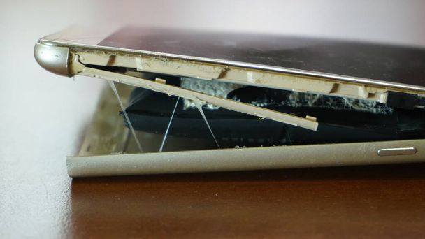 Swollen smartphone battery. Damaged smartphone with a faulty battery. Damaged phone on a wooden background. - Photo, Image