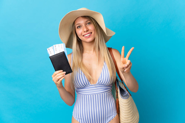 Young blonde woman holding a passport isolated on blue background smiling and showing victory sign - Photo, Image