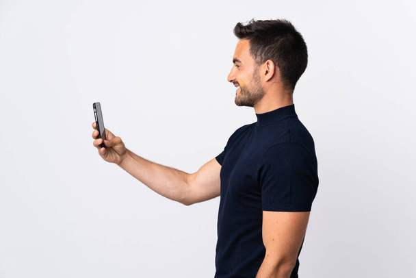 Νεαρός καυκάσιος άνδρας που χρησιμοποιεί κινητό τηλέφωνο απομονωμένο σε λευκό φόντο με χαρούμενη έκφραση - Φωτογραφία, εικόνα