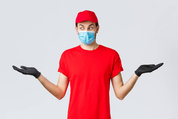 Groceries and packages delivery, covid-19, quarantine and shopping concept. Surprised courier in red cap and t-shirt uniform, wear protective face mask, gloves, hold two items in hands sideways - Photo, image