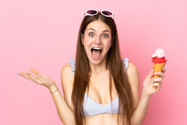 Young caucasian woman in swimsuit holding a cornet ice cream isolated on pink background with shocked facial expression - Foto, afbeelding